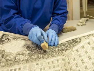 Trockenreinigung Zwei Hände in blauen Handschuhen reinigen mit einem Kautschukschwamm einen Kupferstich mit Gebäudedarstellung.