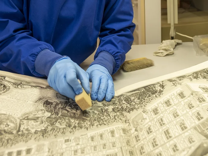 Trockenreinigung Zwei Hände in blauen Handschuhen reinigen mit einem Kautschukschwamm einen Kupferstich mit Gebäudedarstellung.
