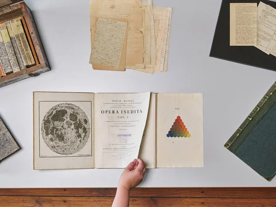Tisch mit historischen Büchern und Archivmaterialien, eine Hand blättert in einem Buch