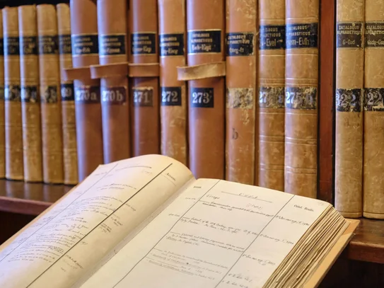 Recherchieren Im Hintergrund Bibliothekskataloge auf einem Regal, im linken vorderen Bereich ein aufgeschlagener Bibliothekskatalog