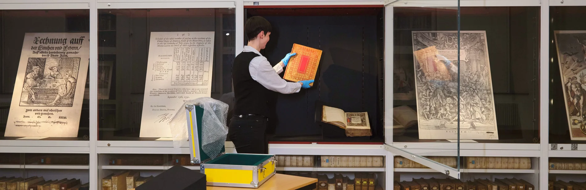 Restauratorin legt ein Buch in eine offene Vitrine; daneben geschlossene Vitrinen mit Faksimiles.
