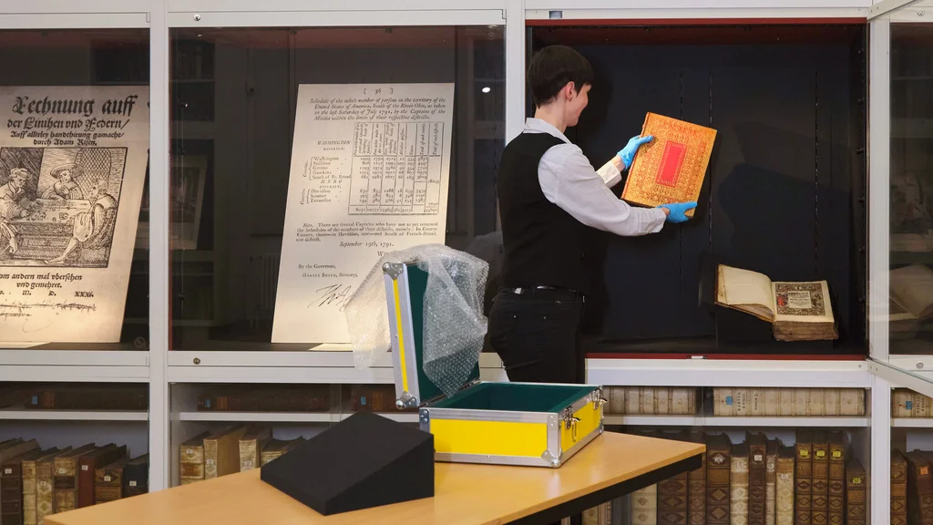 Ausstellungsvorbeitung an der SUB Göttingen Restauratorin legt ein Buch in eine offene Vitrine; daneben geschlossene Vitrinen mit Faksimiles.
