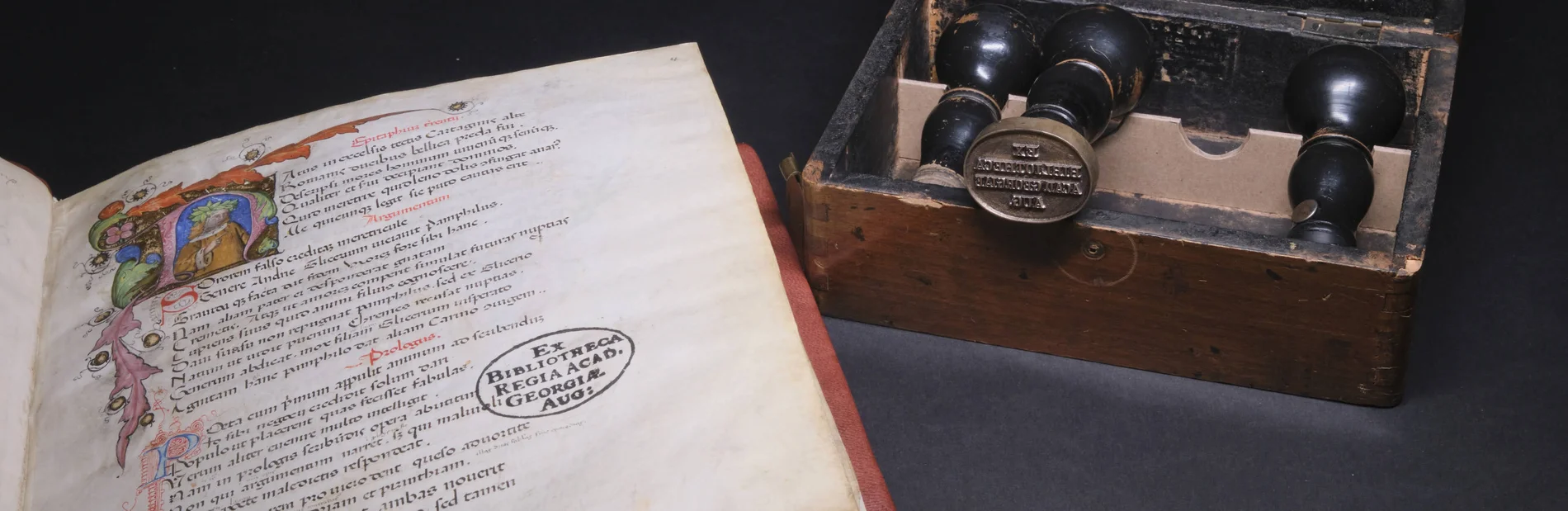 Links eine aufgeschlagene Handschrift mit dem Stempel „EX BIBLIOTHECA REGIA ACAD. REGIAE AUG:“, rechts ein Holzkasten mit historischen Stempelwerkzeugen.