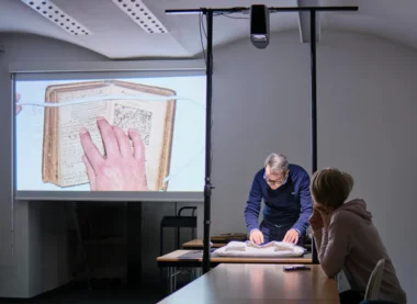 Mann beugt sich über ein altes Buch, dessen Inhalt an die Wand projiziert wird. Im Vordergrund rechts sitzt eine Frau und betrachtet den Bildschirm.