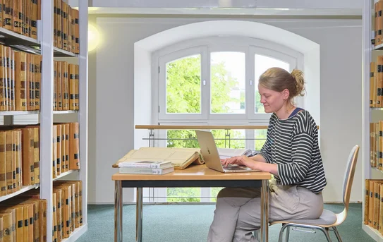 Besuch im Lesesaal Nutzerin sitzt an einem Tisch zwischen Bücherregalen und tippt auf einem Laptop.