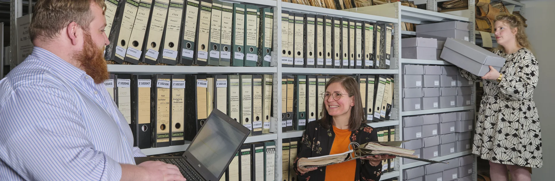 Drei Personen vor einem Regal mit Archivmaterial, eine Person nimmt eine Box heraus.