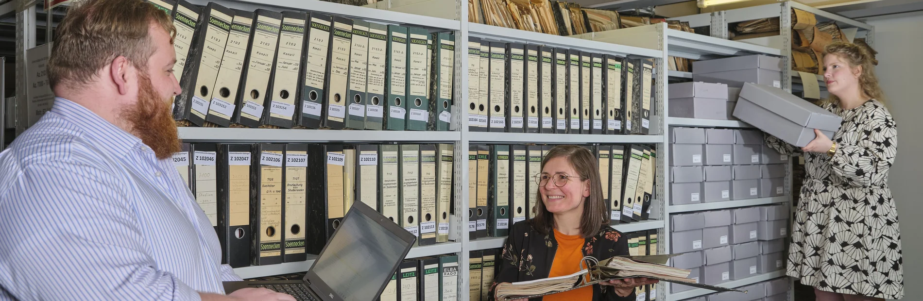 Arbeiten im Universitätsarchiv Drei Personen vor einem Regal mit Archivmaterial, eine Person nimmt eine Box heraus.